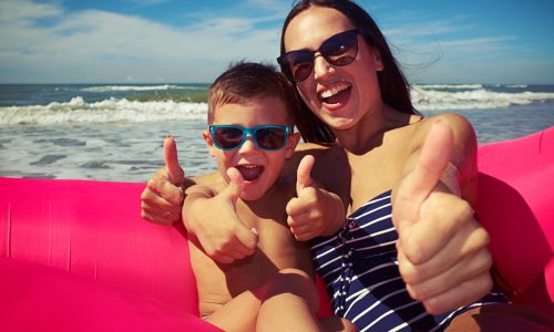 Mid shot of the happy family who are sitting on the rubber raft on the beach and holding their thumbs-up on a gorgeous sunny day. Enjoying time together